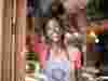 Happy waitress wearing pink t shirt and grey apron, smiling at camera with restaurant behind her